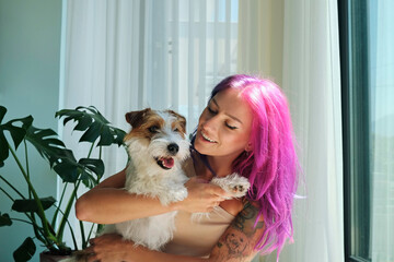 Wall Mural - Young beautiful woman with pink hair hugging with her wire haired jack russell terrier at home. Broken coated pup in arms of smiling female. Close up, copy space, background.