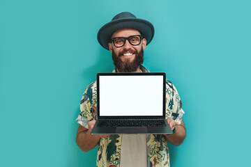 Portrait of a cheerful hipster guy with a beard in a Hawaiian shirt holding a laptop, showing blank screen with copy space for advertisement and promotional text, demonstrates something.