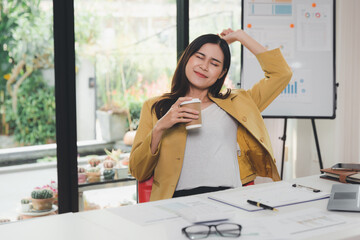 A woman holding a cup of coffee and stretch lazily relax from fatigue after work all day in home office. sleepy, tired, overworked, lazy to work. Asian businesswoman stretching her arms and relaxing.