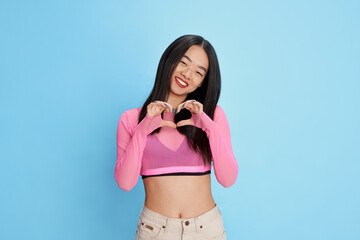 Wall Mural - Positivity. Portrait of young beautiful smiling girl showing heart gesture with hands, posing against blue studio background. Concept of emotions, facial expression, fashion, youth, lifestyle. Ad