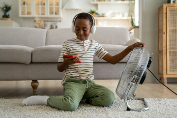 Wall Mural - Happy African American child schoolboy enjoying free leisure time after school at home, sitting on floor in front of electric fan, holding smartphone playing online game. Summer heat and kids