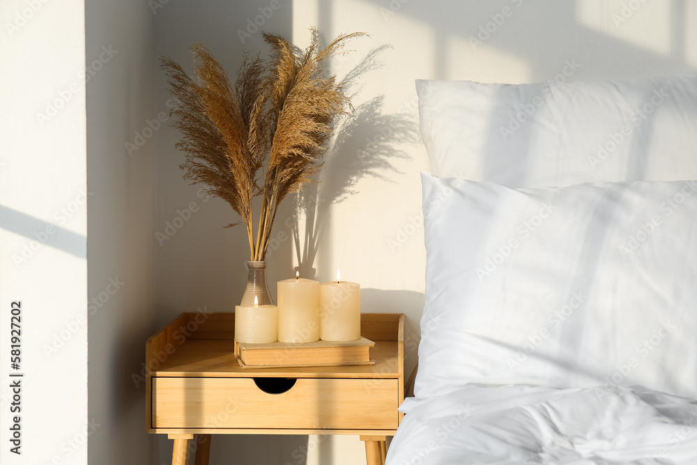 Burning candles, book and vase with pampas grass on table near light wall