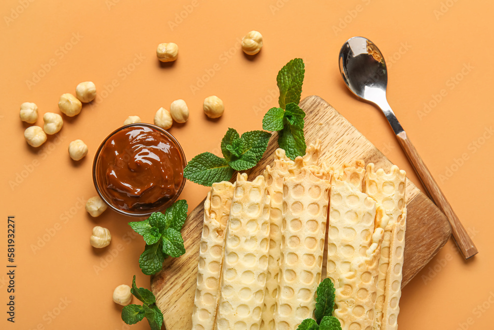 Board with delicious wafer rolls, boiled condensed milk, mint and hazelnuts on orange background
