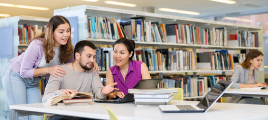 Cheerful young adult university students studying in library using books and laptop, talking and smiling while working on group project. Brainstorming and teamwork concept