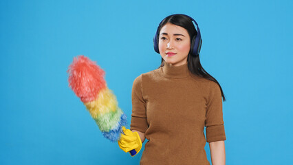 Wall Mural - Cheeful housekeeper wearing headphones while dancing and having fun during house cleaning using feather duster. Overwhelmed maid role was vital to the smooth operation and cleanliness of many homes