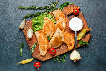 wooden board with tasty meat empanadas, vegetables, herbs and sauce on dark background