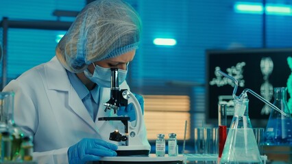 Wall Mural - A female scientist examines a sample under a microscope. A woman doctor in a white medical gown, bonnet, blue gloves and a mask. Laboratory with test tubes, flasks and computer monitors. Close up.