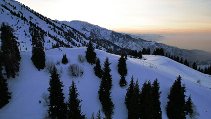 Wall Mural - Epic red-orange sunset in high snowy mountains. There are tall coniferous trees, there is snow on the hills. High peaks. The sun's rays fall on the clouds and fog that is lower down the gorge. Almaty