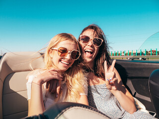 Portrait of two young beautiful and smiling hipster female in convertible car. Sexy carefree women driving cabriolet. Positive models riding and having fun in sunglasses outdoors. Enjoying summer days