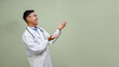 © bongkarn - Laughing Asian male doctor in white gown is looking aside, opening his palm at the empty space