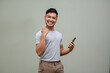 © bongkarn - Cheerful Asian man showing thumb up hand gesture, holding his phone and smiling to camera