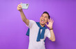 © Bangun Stock Photo - Cheerful young Asian man in white t-shirt doing selfie shot on mobile phone and waving hand isolated on purple background