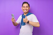 © Bangun Stock Photo - Cheerful young Asian man using mobile phone and showing thumb up gesture isolated over purple background