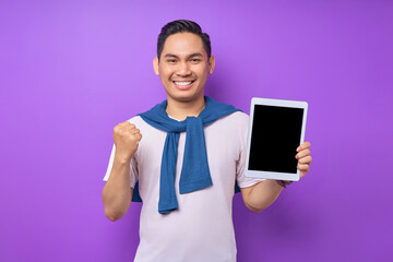 Excited young Asian man in casual clothes showing digital tablet with empty screen for mockup and celebrating success isolated over purple background