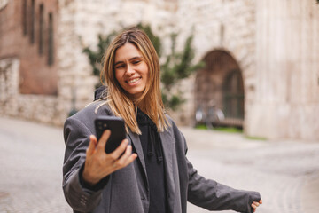 Wall Mural - Happy young woman photographing herself using her mobile phone. Caucasian female talking selfie with her smart phone walking outdoor on the street. Girl posing in grey suit, t-shirt.