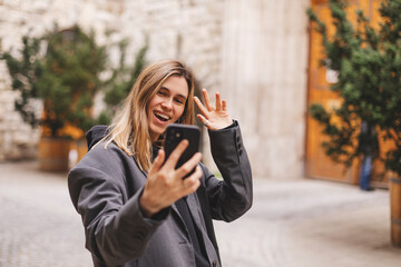 Wall Mural - Woman waiving saying hello happy and smiling, friendly welcome gesture. Portrait of young beautiful attractive smiling cheerful girl taking selfie saying hello outside outdoors.