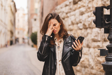 Sad woman checking bad news on mobile phone complaining alone in the street. Upset young woman thinking about her problems. Girl cover her face by her hand. Negative, bad news, breakup, unhappy.