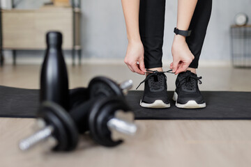 sport and healthy lifestyle concept - woman preparing for training at home and tying shoelaces on sneakers - dumbbells and bottle of water on first plan