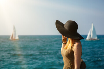 Silhouette of a girl on the background of the blue sea and blue sky. sailing yachts sail across the sea and the model looks in their direction. The black hat with blond hair visible under