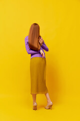 Faceless portrait of slim woman with long straight red hair wearing retro fashion outfit in action over yellow studio background. Impersonal emotions, body language