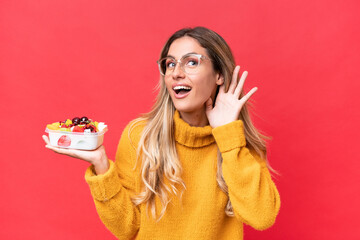 Canvas Print - Young pretty Uruguayan woman holding a bowl of fruit isolated on red background listening to something by putting hand on the ear