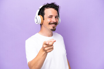 Wall Mural - Young caucasian man isolated on purple background listening music and pointing to the front