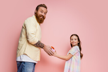 Wall Mural - Cheerful bearded father and preteen girl holding gift box and looking at camera on pink background.