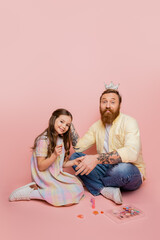 Wall Mural - Positive girl holding nail polish near tattooed dad with crown on head and decorative cosmetics on pink background.