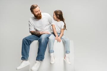 Wall Mural - Positive tattooed man looking at daughter in t-shirt and jeans sitting on cube isolated on grey.