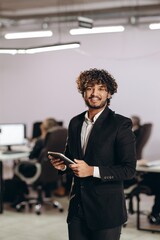 Wall Mural - Handsome young businessman with digital tablet. Indoor shot of curly office worker in suit using device