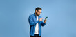 © Moon Safari - Serious handsome young adult man dressed in denim shirt surfing internet over smart phone while standing isolated against blue background