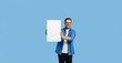 © Moon Safari - Young adult smiling businessman entrepreneur wearing denim shirt and eyeglasses showing white blank poster for marketing isolated on blue background