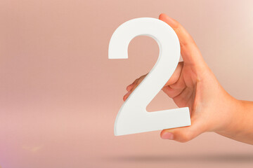 Fototapete - Number two in hand. Hand holding white number 2 on red background with copy space. Concept with number two. 2 percent, birthday 2 years old, couple, two, double