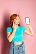 © Georgi - A happy red-haired young girl is showing a blank white smartphone for a copy space and advertisement against an isolated pink background.