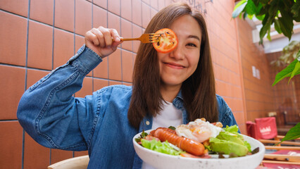 Wall Mural - Portrait image of a young woman playing and covering eye with a piece of tomato while eating salad