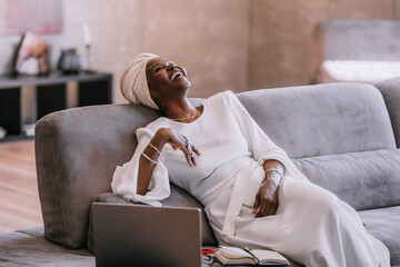 Excited African young woman in white turban and traditional dress relaxing on couch at home laughing out loud after friends joke during distant video call using laptop. Happy people, business, finance