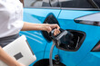© ND STOCK - Woman plugging in the charger into a socket of her blue electric car at a charging station in the street