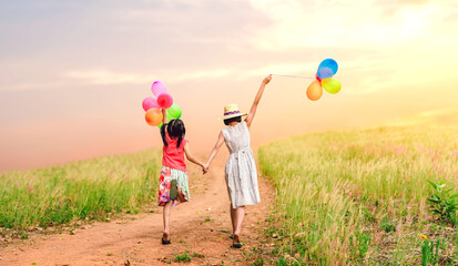 Wall Mural - Happy Two children girls holding balloons walking on meadow trail springfield sunset background.