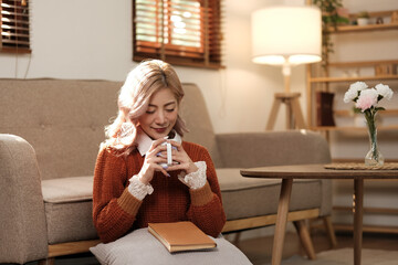 Image of stylish young woman in modern house, using mobile phone, sitting on floor and holding smartphone, drinking from cup