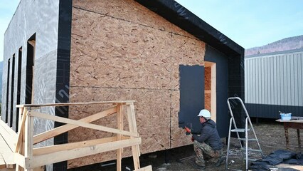 Canvas Print - Male painter using paint roller, doing exterior paint work in a black color. Man worker building wooden frame house. Carpentry and construction concept.