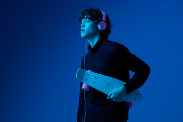Wall Mural - Teenage male with headphones listening to music and dancing with skateboard in hand, hipster lifestyle, portrait dark blue background, neon light, style and trends, mixed light, copying space