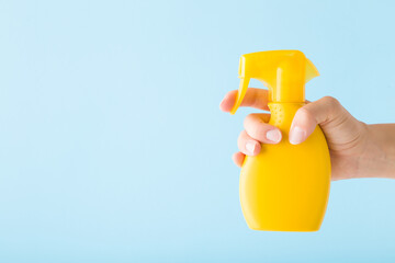 Wall Mural - Young adult woman hand holding yellow spray bottle of sunscreen or cleaning detergent on light blue wall background. Pastel color. Closeup. Empty place for text. Side view.