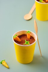 Wall Mural - Dessert, carrot cake to go  in a yellow paper cup, decorated with cocoa and marzipan carrots on a green plain background. Trendy cakes.