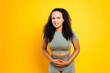 © Kateryna - Unhealthy brazilian or hispanic young woman in sportswear, keeps hands on a stomach, demonstrating abdominal pain, pain during menstruation, looks sadly at camera, grimacing,isolated yellow background