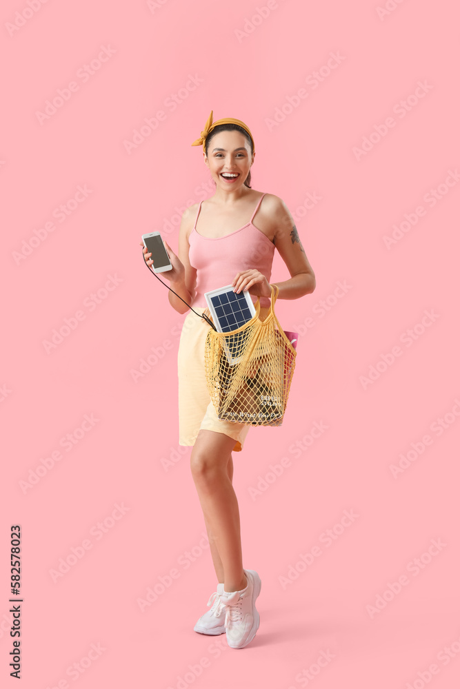 Pretty young woman charging mobile phone with portable solar panel on pink background