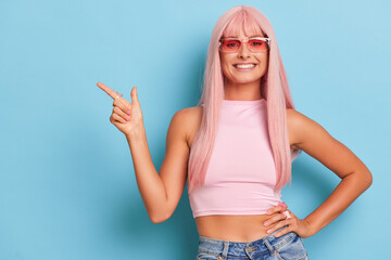 pleasant model with pink hair wearing pink sunglasses and white top standing against blue background