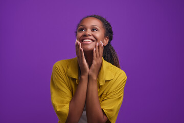 Wall Mural - Young happy excited African American woman touching chin and leaning down delighted with invitation to summer trip from boyfriend or hearing other good news standing posing on purple background