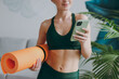© ViDi Studio - Close up young sporty athletic fitness trainer instructor woman wearing green tracksuit hold in hand yoga mat use mobile cell phone training do exercises at home gym. Workout sport motivation concept.