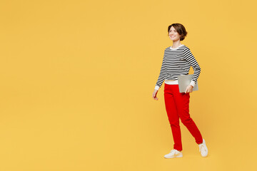 Full body side view young woman wear casual black and white shirt hold closed laptop pc computer walking going strolling isolated on plain yellow color background studio portrait. Lifestyle concept.