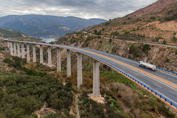 Canvas Print - Landscape with a very long and high viaduct in the foreground that surrounds a reservoir, and where a fuel tanker truck circulates, top view.